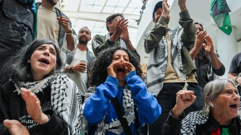 A group of people wearing black-and-white keffiyeh scarves chant and raise their hands inside a building with a glass skylight ceiling.