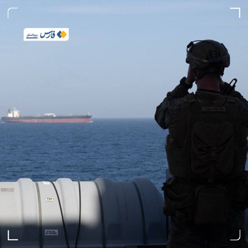 A soldier in camouflage gear with a backpack labeled "CHAMBERS" looks through binoculars from a ship's deck toward a large tanker on the open sea.