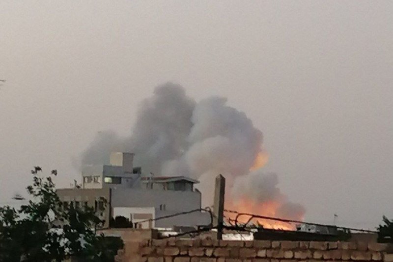 Flames and a large plume of dark smoke rise behind white buildings in a residential area, with a brick wall and barbed wire visible in the foreground.