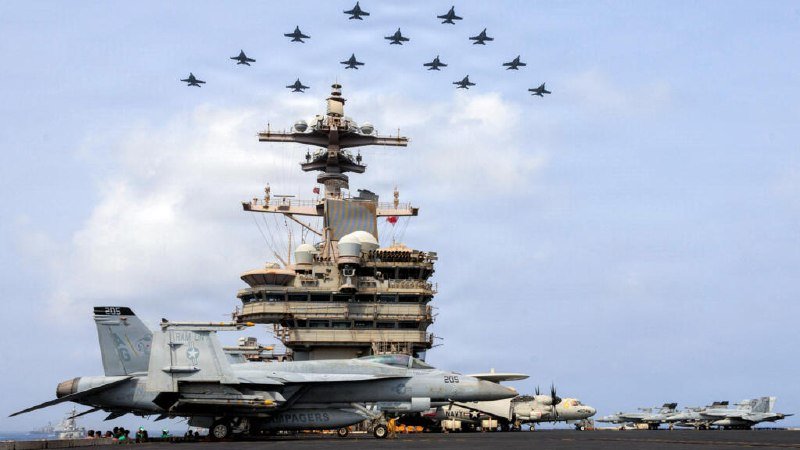 A formation of fighter jets flies in a diamond pattern above an aircraft carrier's tower, while another jet marked "205" sits on the flight deck.