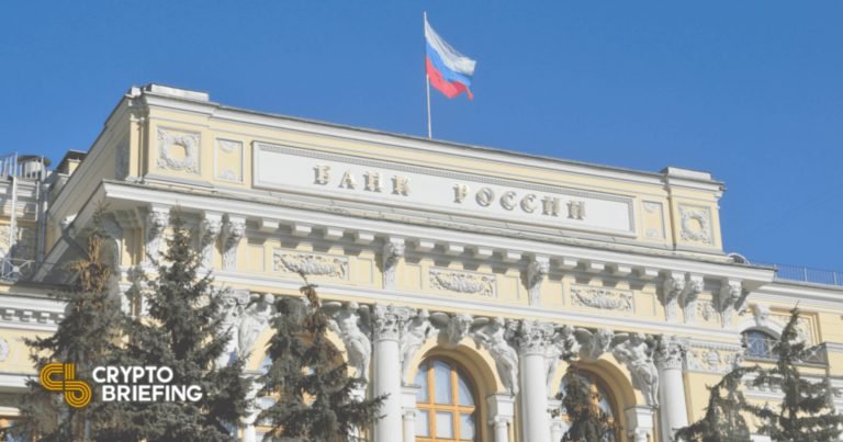 The Bank of Russia building in Moscow, featuring classical architecture with columns, ornate yellow facade, and the Russian tricolor flag flying above under a clear blue sky.