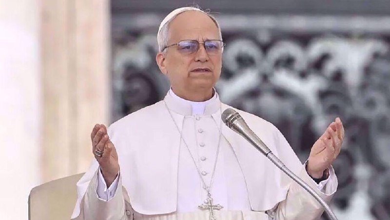 A clergyman in white robes and a zucchetto speaks into a microphone with both hands raised.