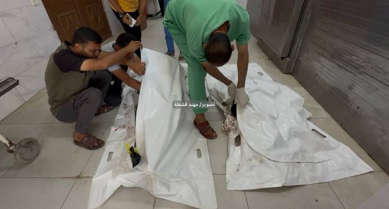 Men kneel on a tiled floor, pulling back white body bags stained with blood to reveal the remains inside.