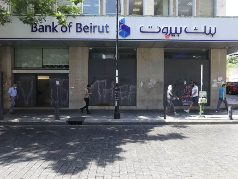Pedestrians walk past a Bank of Beirut branch with graffiti-covered walls and shuttered doors on a sunny street.