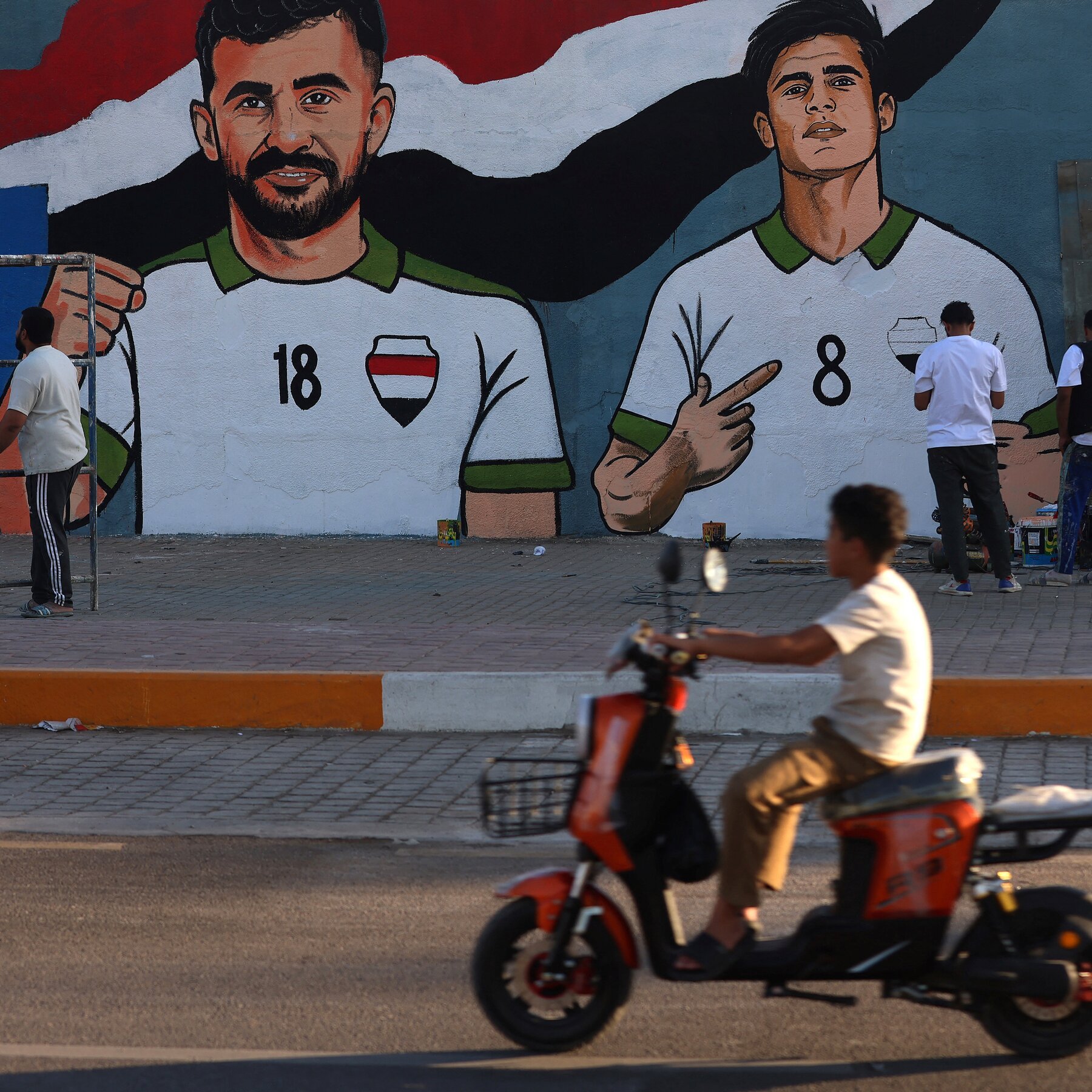 A person rides an orange scooter past a large wall mural depicting two men in white and green jerseys numbered 18 and 8.