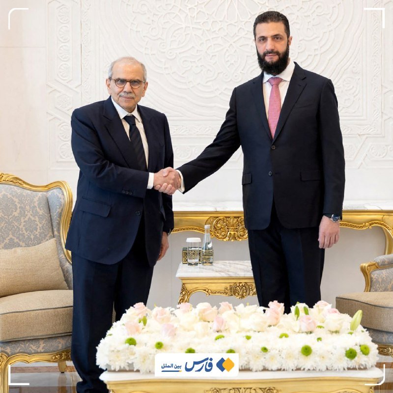 Two men in dark suits shake hands in an ornately decorated room with white carved walls, gold-trimmed furniture, and a floral arrangement in the foreground.