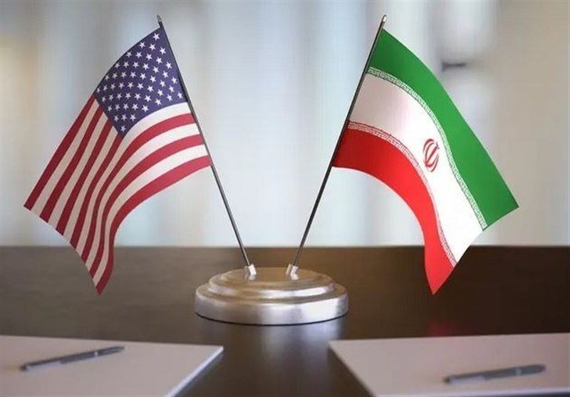 Small American and Iranian flags stand crossed on a shared base atop a dark wooden desk, with two notebooks and pens placed nearby.