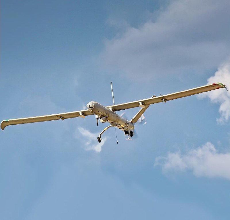 A military drone flies against a blue sky with scattered white clouds.