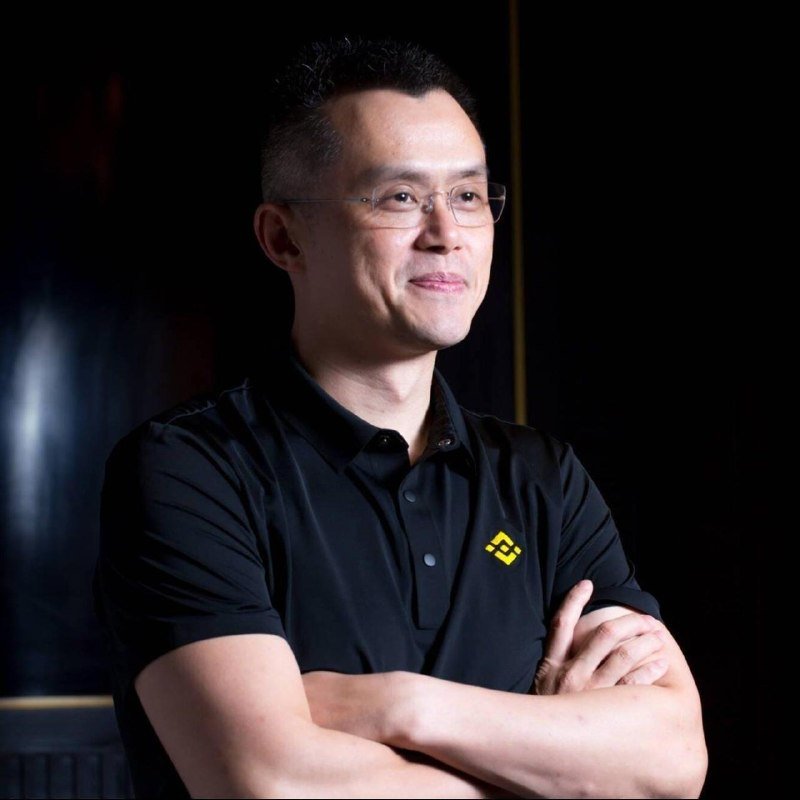 A man wearing glasses and a black polo shirt with a yellow logo stands with crossed arms against a dark background.