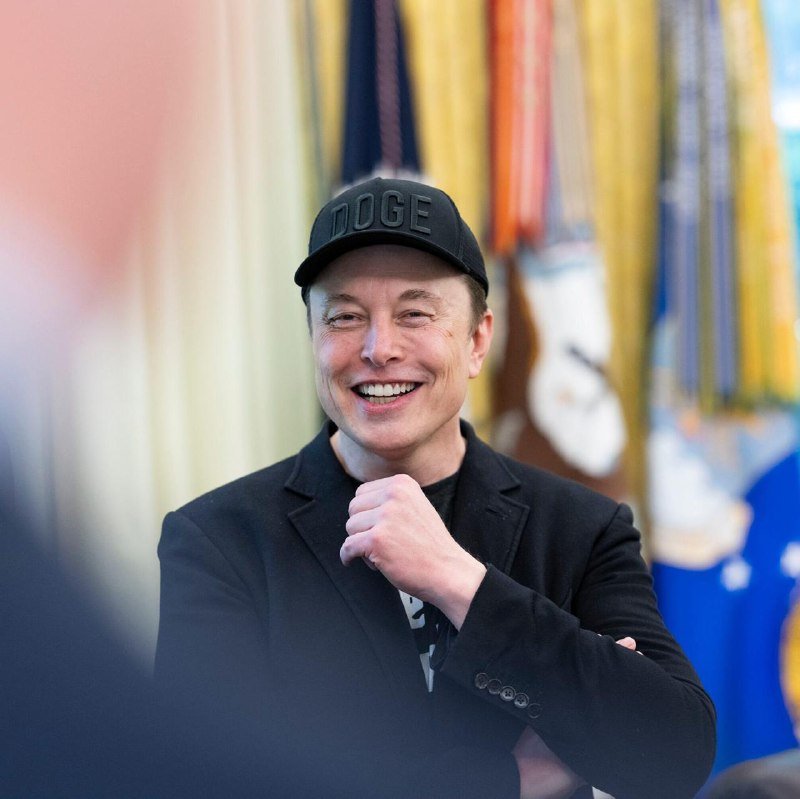 A smiling man wearing a black "DOGE" cap and dark blazer sits in front of several flags, his fist raised near his chin.