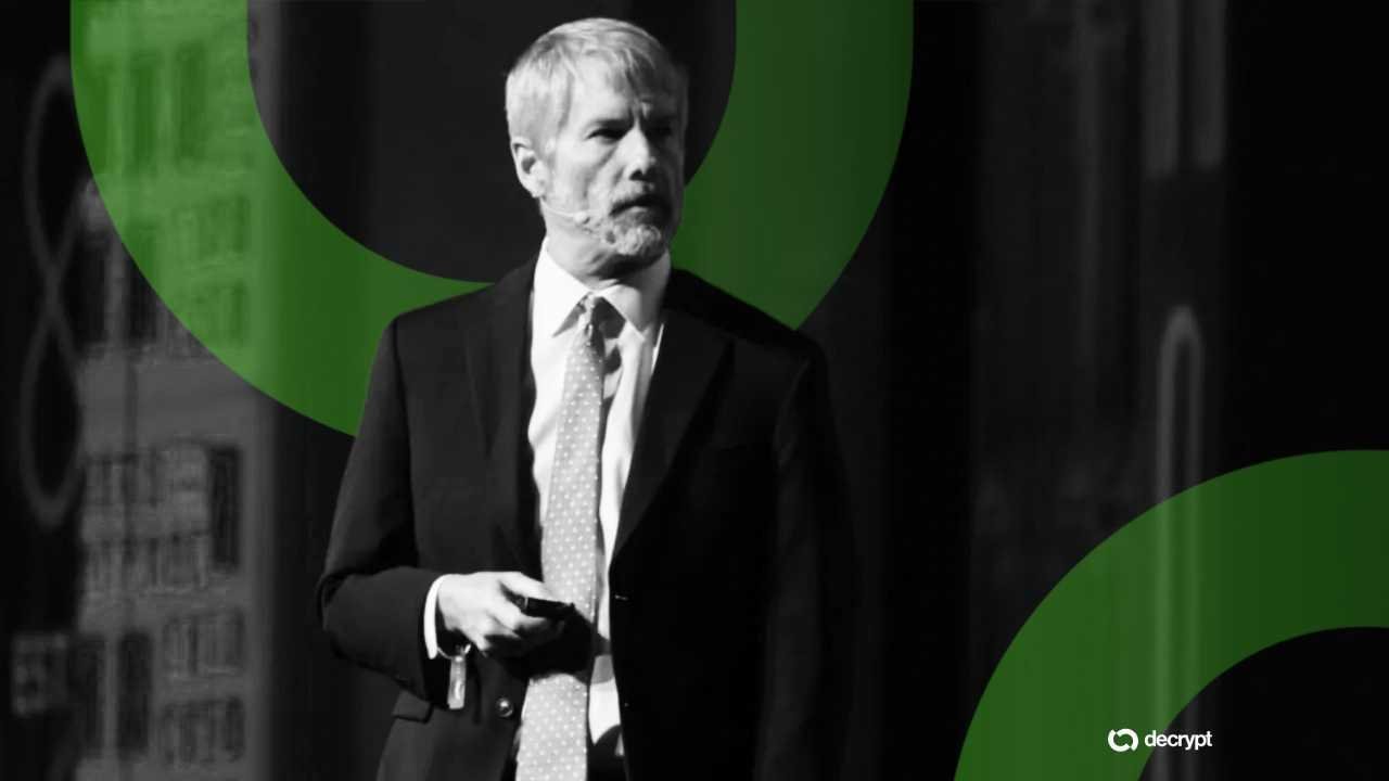 A gray-haired man in a dark suit and tie stands on stage against a dark backdrop with green graphic elements and a "decrypt" logo.
