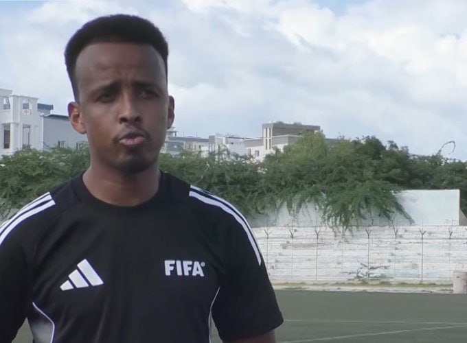 A man wearing a black Adidas FIFA shirt stands on an outdoor sports field, with a wall topped with barbed wire and buildings visible in the background.