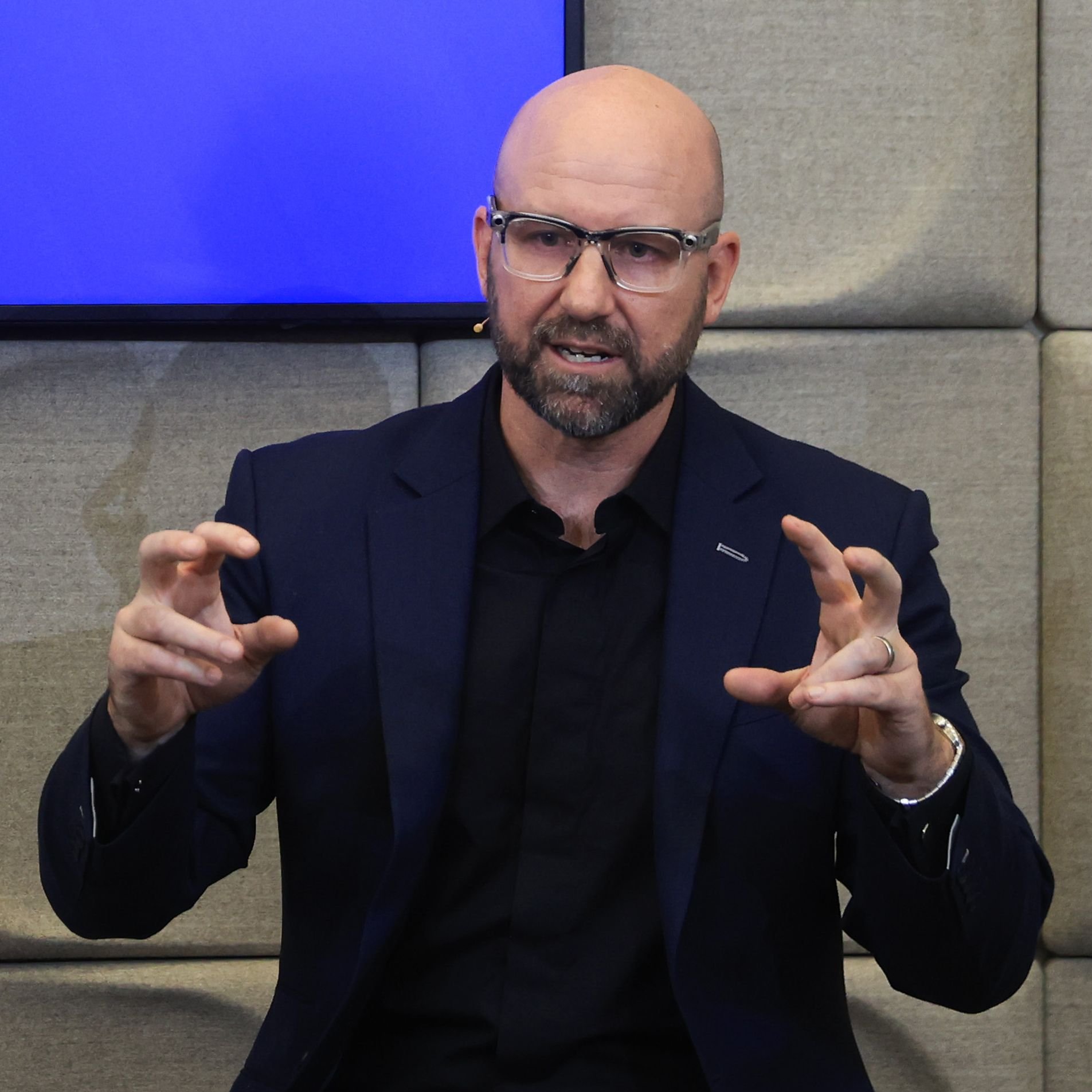 A bald, bearded man in a dark blazer and clear-framed glasses gestures with both hands while speaking in front of a paneled wall and screen.