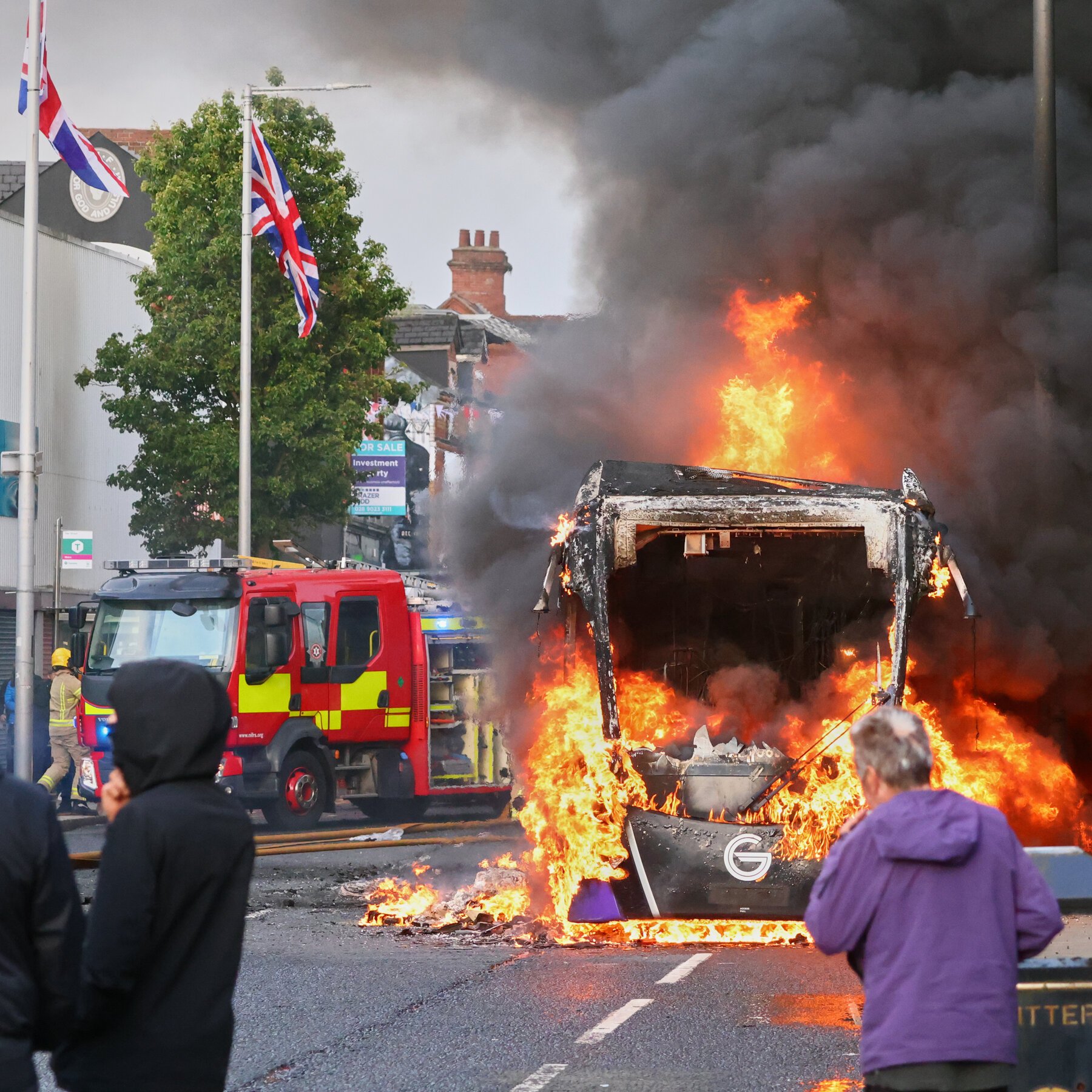 A bus engulfed in flames and thick black smoke on a street, with a red fire engine, a firefighter, and bystanders nearby, and Union Jack flags flying on poles.