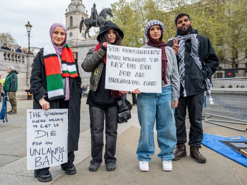 Four people stand in a public square holding signs reading "The government were willing to let us die for an unlawful ban," with an equestrian statue visible in the background.