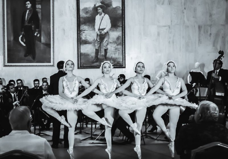 Four ballerinas in white tutus perform in synchronized pose before an orchestra, with two large framed portraits hanging on the wall behind them.
