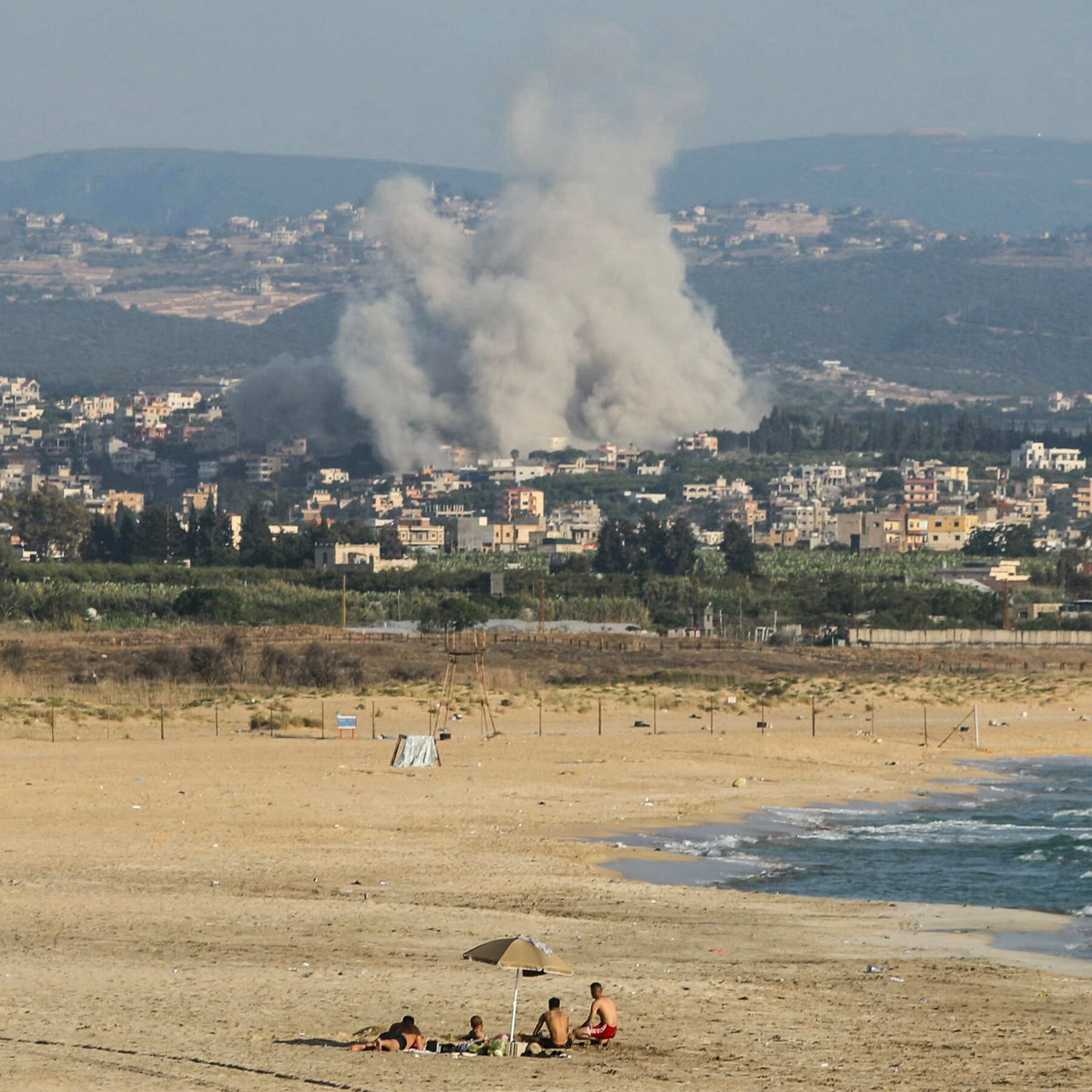 People sit under a beach umbrella on a sandy shore as large plumes of smoke rise from a town visible in the background.