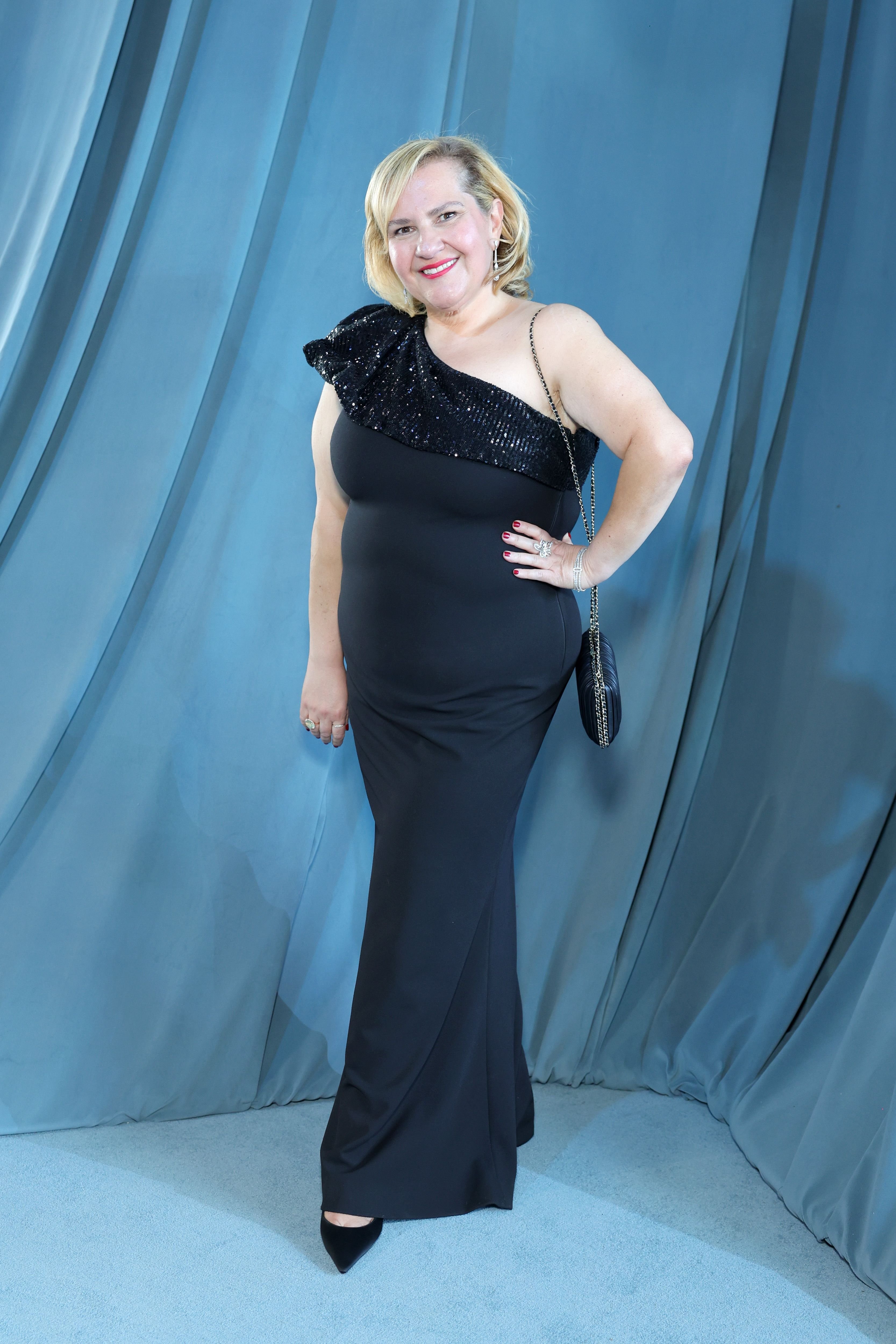 A smiling woman with blonde hair poses in a one-shoulder black sequined gown with a quilted chain-strap handbag, standing against a draped blue-gray backdrop.