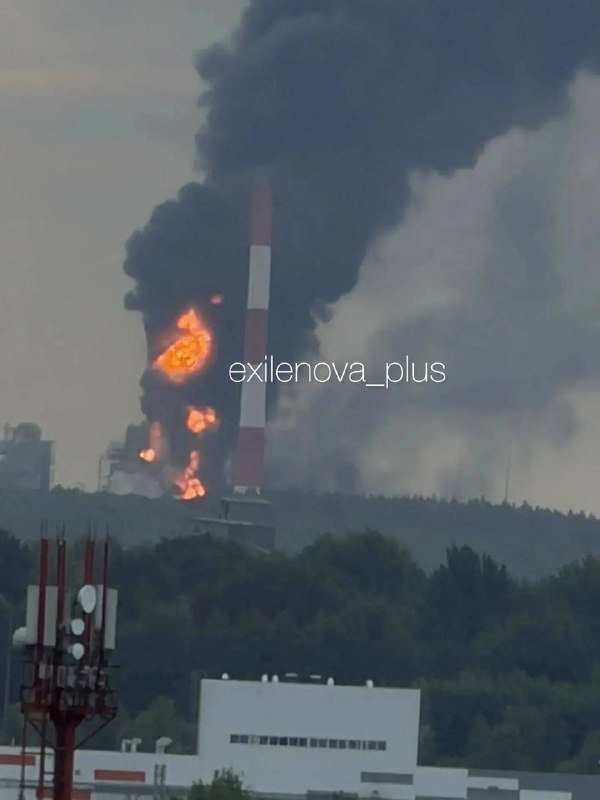 A red and white industrial smokestack is engulfed in flames, with thick black smoke billowing into the sky above nearby trees and buildings.
