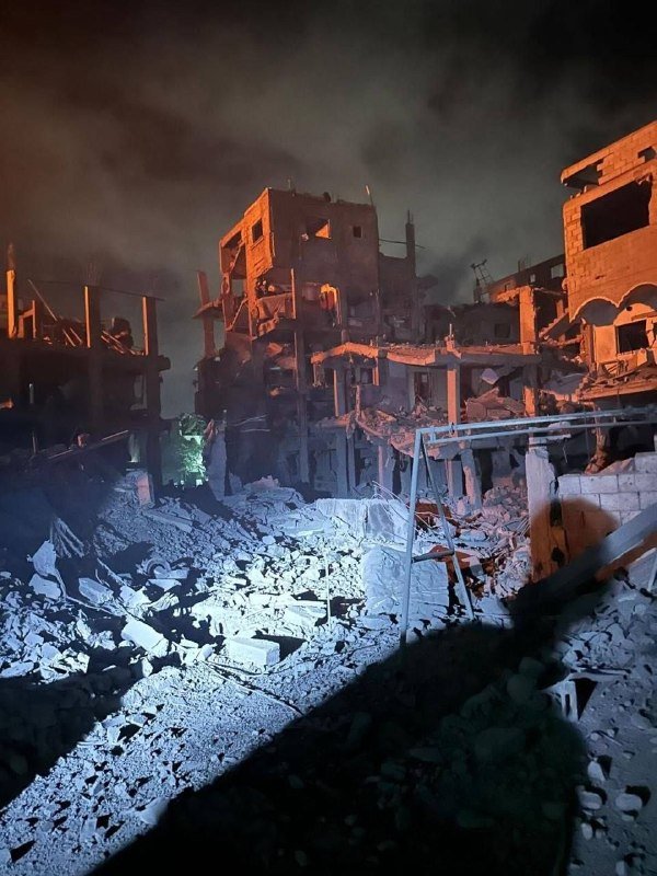 Heavily damaged and collapsed concrete buildings stand in rubble at night, illuminated against a dark sky.