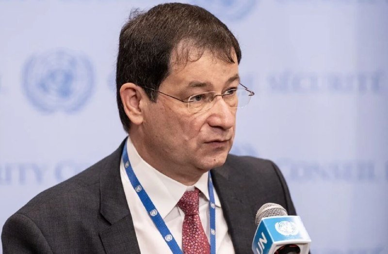 A bespectacled man in a dark suit and red tie speaks into a UN-branded microphone, with a blue UN emblem backdrop behind him.
