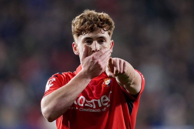 A curly-haired soccer player in a red jersey with "Osasuna" branding kisses his hand and points forward while celebrating, set against a blurred stadium crowd.