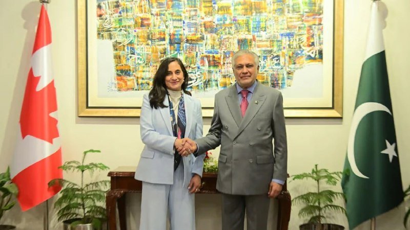 A woman in a light blue suit and a man in a grey suit shake hands in front of the Canadian and Pakistani flags and a large framed abstract painting.
