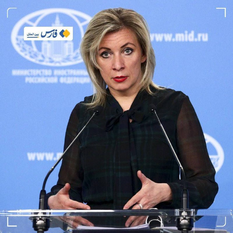 A blonde woman in a dark plaid blouse speaks at a podium with two microphones, gesturing with her hands, against a blue backdrop displaying the URL "www.mid.ru."