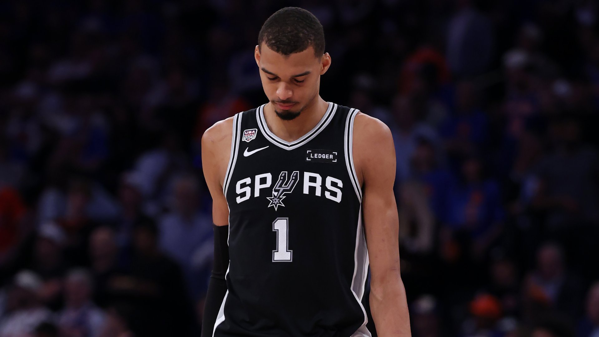A basketball player wearing a black San Antonio Spurs jersey with the number 1 looks downward during a game, with a blurred crowd visible in the background.