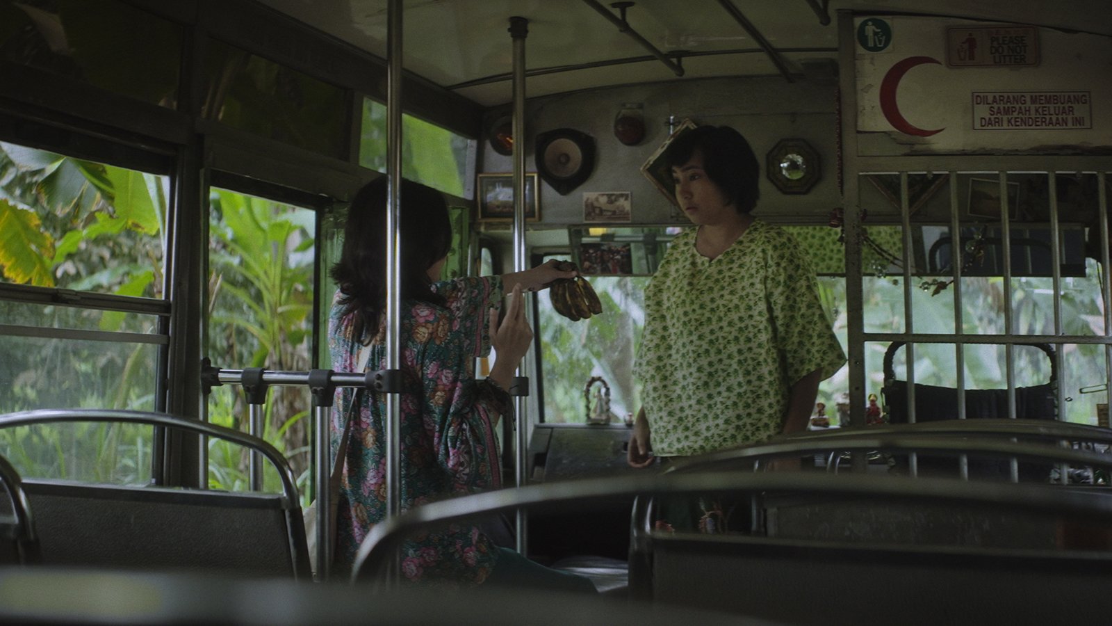 Two women stand inside a bus, one in a floral dress holding a small object toward the other, who wears a green patterned top, with a "Please Do Not Litter" sign visible above.