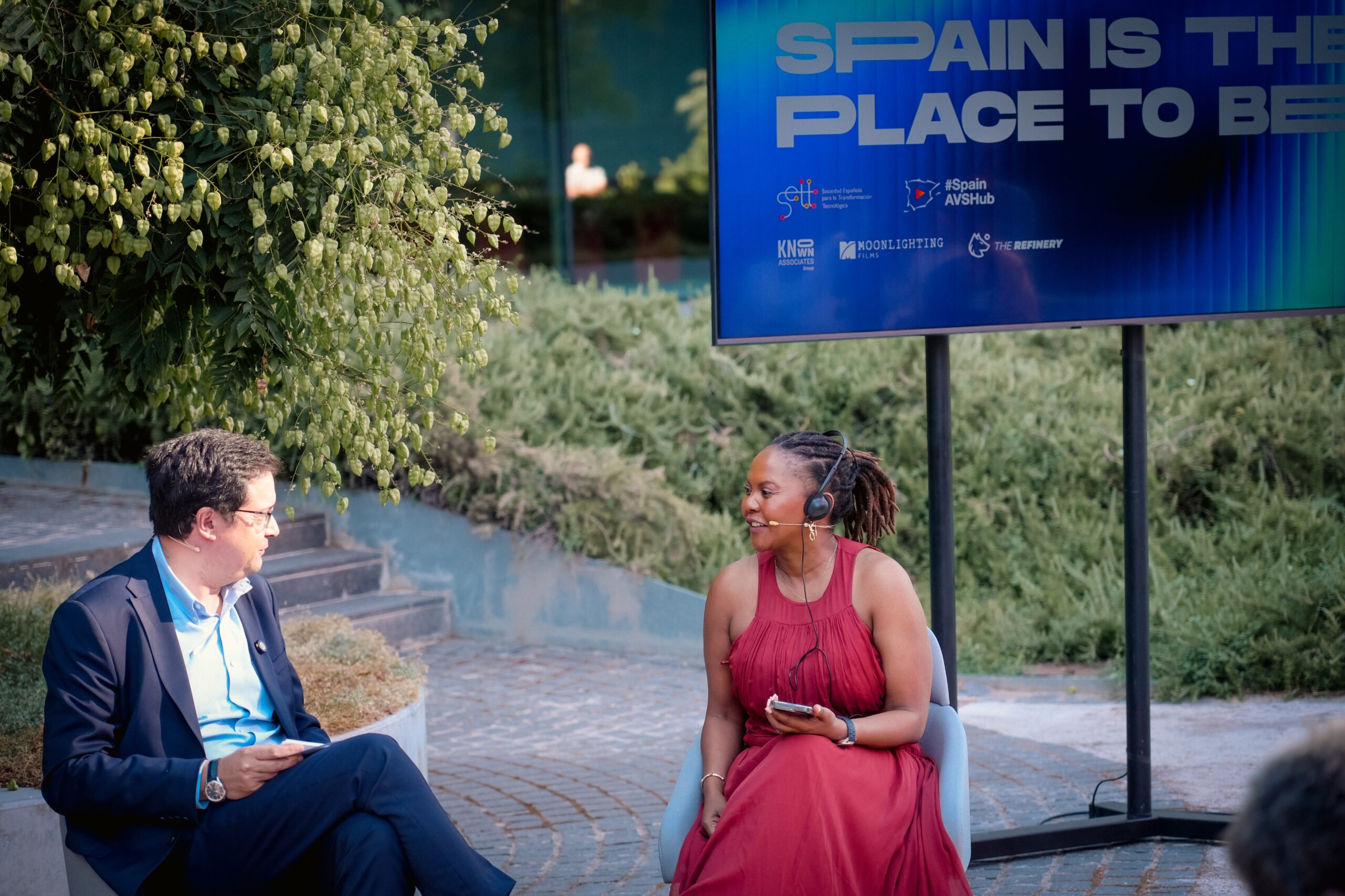 A man in a dark suit and a woman in a red dress with a headset converse while seated outdoors near a screen displaying "SPAIN IS THE PLACE TO BE."