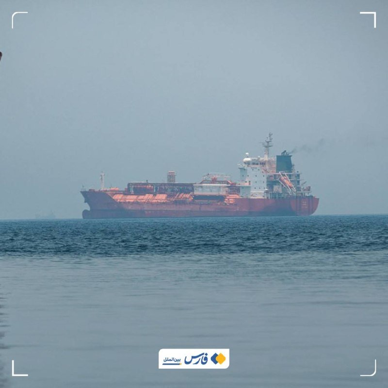 A large red and black cargo tanker ship sails on calm open sea waters under a hazy gray sky.