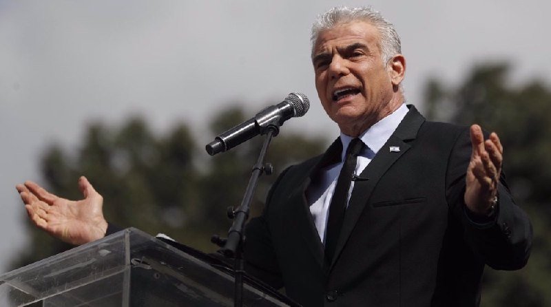 A gray-haired man in a dark suit and tie stands at a clear podium, speaking into a microphone with both hands raised.