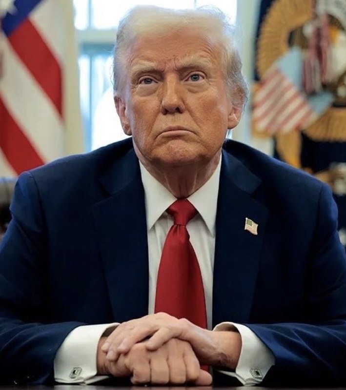An older man with blonde hair in a navy suit and red tie sits with hands clasped, with an American flag and presidential seal visible behind him.