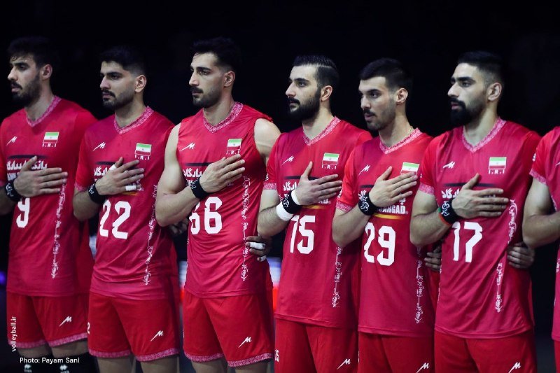 Seven athletes in red jerseys stand in a row with their hands over their hearts and arms around each other.