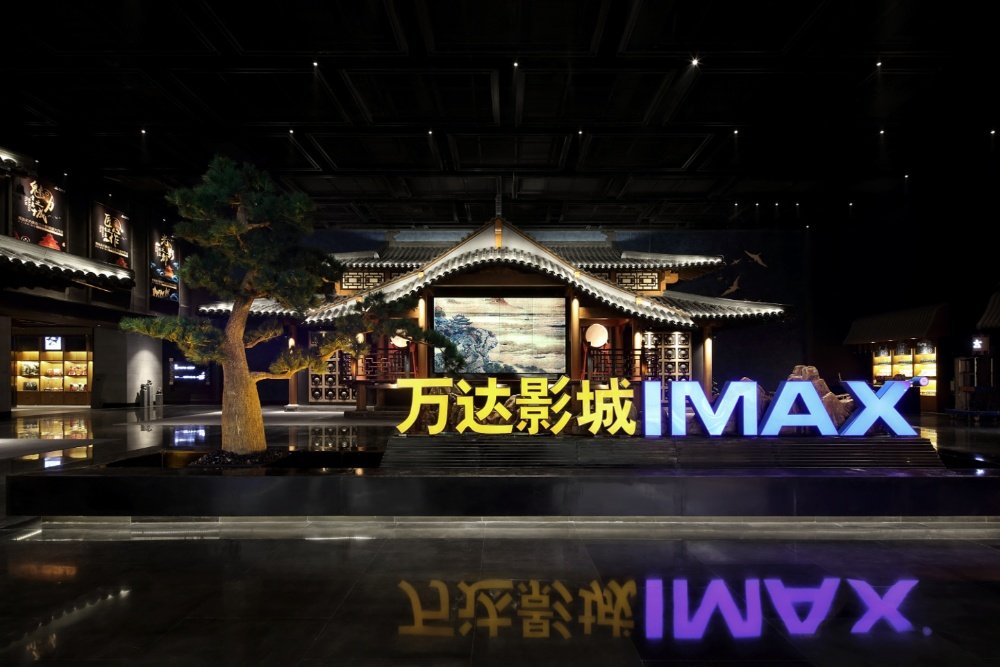 A traditional Chinese pavilion-style structure stands in a dimly lit cinema lobby, with a glowing "万达影城 IMAX" sign and a large pine tree in the foreground.