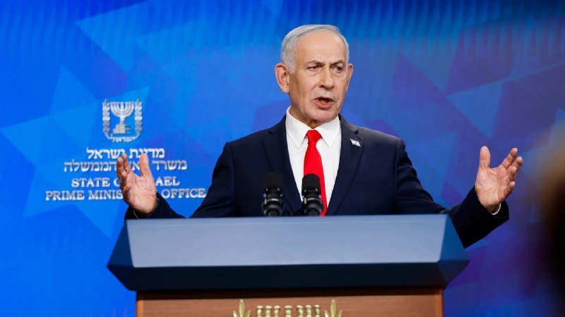 A man in a dark suit and red tie speaks at a podium with raised hands, against a blue backdrop bearing the State of Israel Prime Minister's Office emblem.