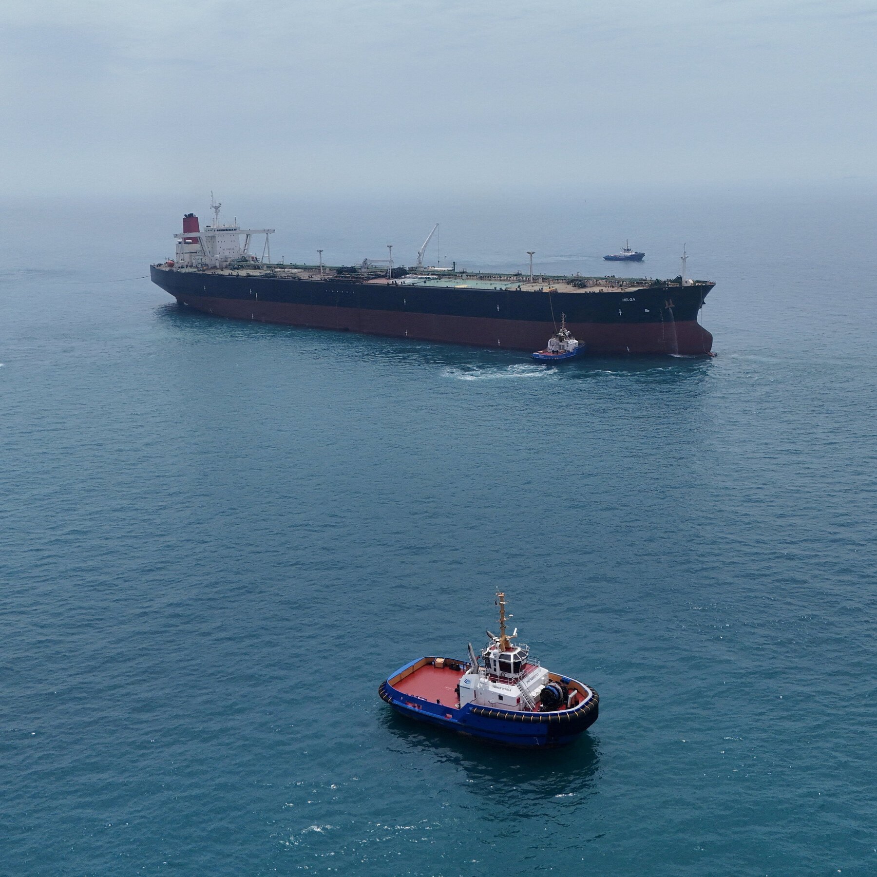 An aerial view shows a large tanker ship labeled "HELGA" accompanied by smaller tugboats on calm open water.