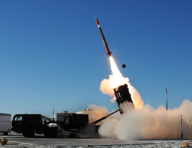 A missile launches skyward from a mobile military launcher truck, with flames and smoke billowing against a clear blue sky.