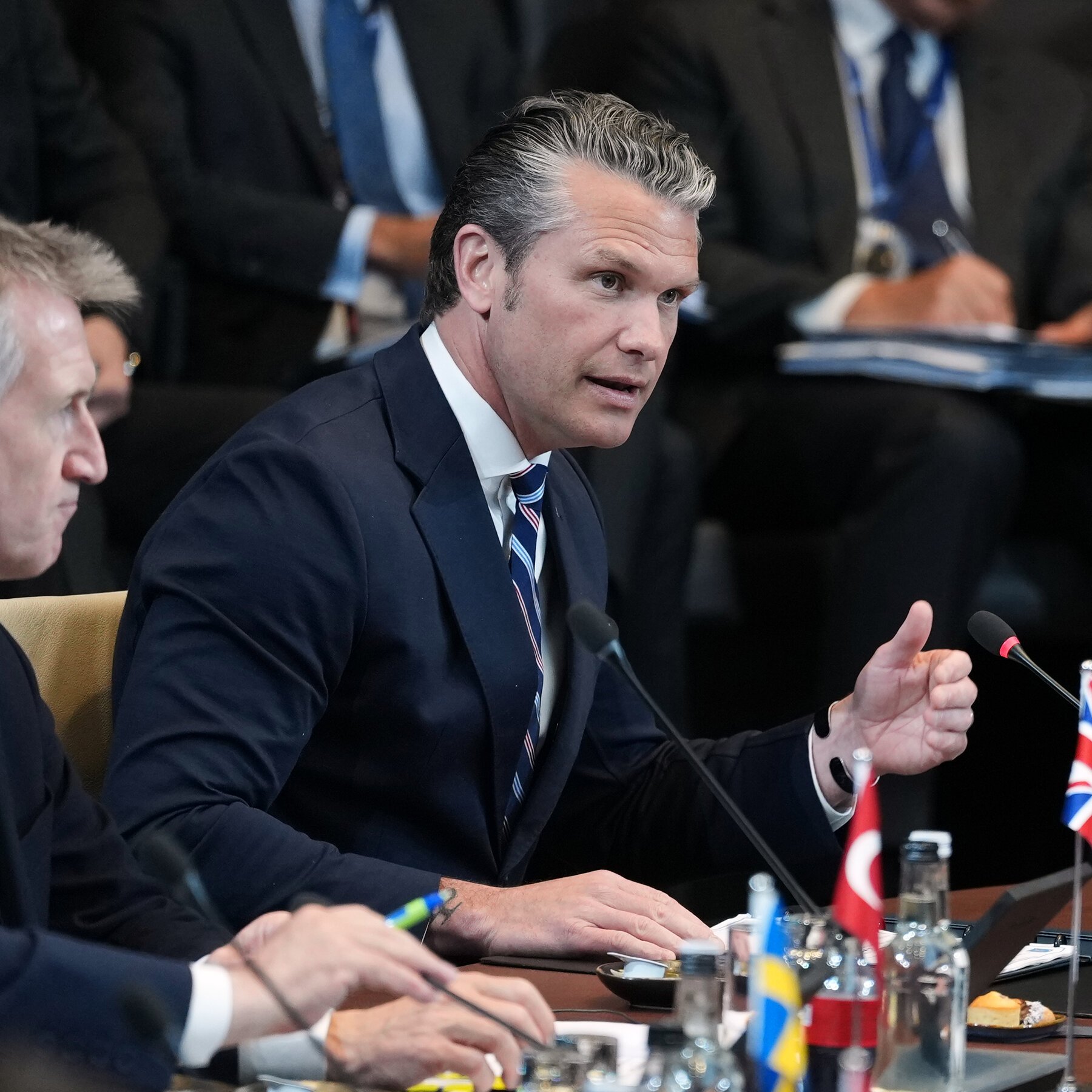 A gray-haired man in a dark suit and striped tie speaks while gesturing with his hand at a table with microphones, small national flags, and water bottles.