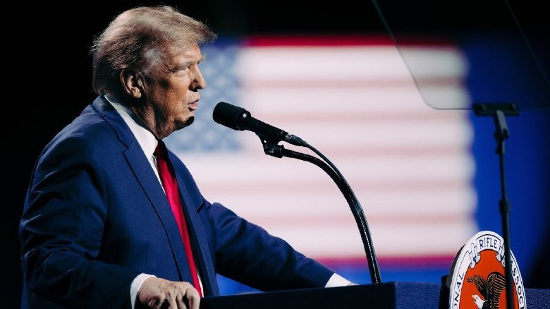 A man in a blue suit and red tie speaks at a podium with a microphone, an American flag and a Marine Corps emblem visible behind him.