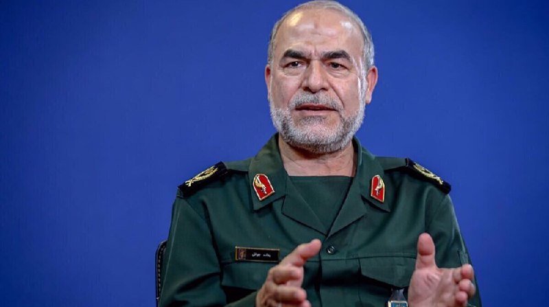 A gray-bearded man in a green military uniform with insignia gestures with both hands against a solid blue background.