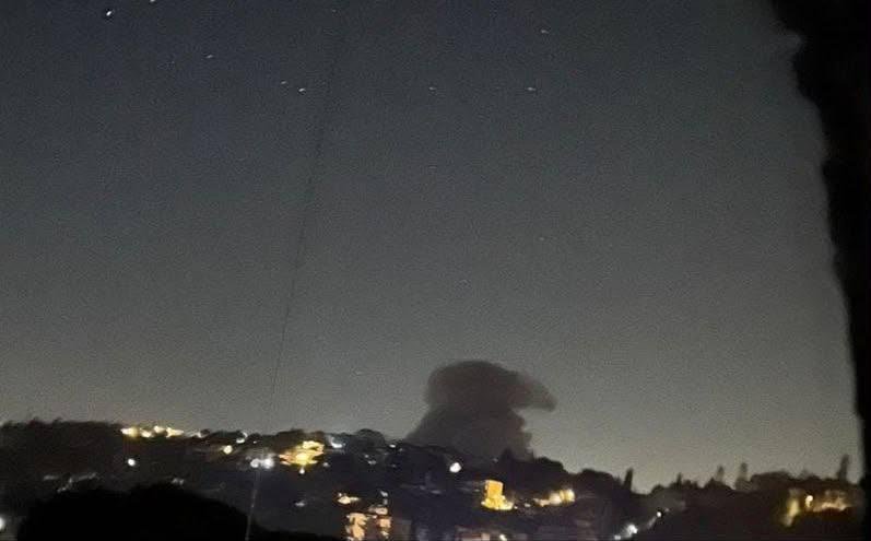 A dark plume of smoke rises above illuminated hillside buildings at night.