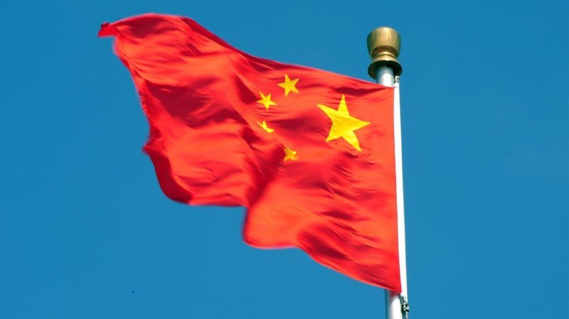 A red flag with one large and four small yellow stars waves against a clear blue sky on a metal flagpole.