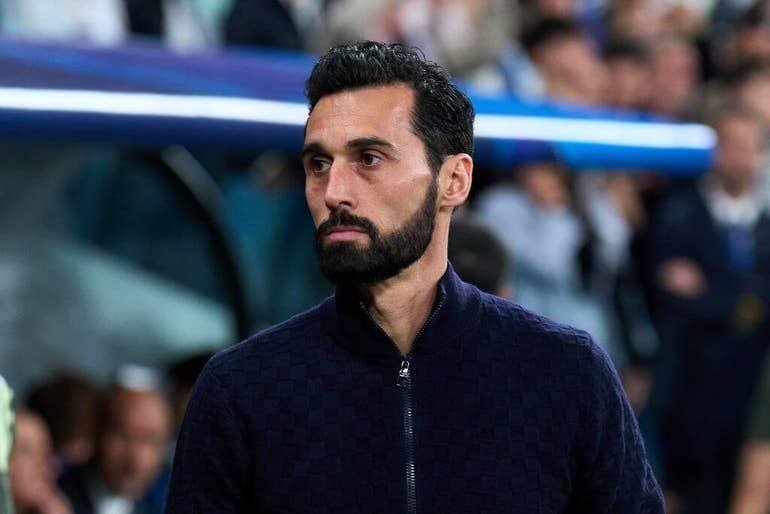 A bearded man in a navy zip-up jacket stands near a stadium dugout, looking off to the side with a serious expression.