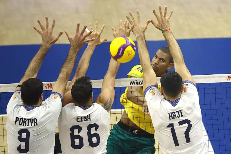 A volleyball player in a yellow jersey spikes a yellow and purple ball over the net against a triple block by defenders in white jerseys.