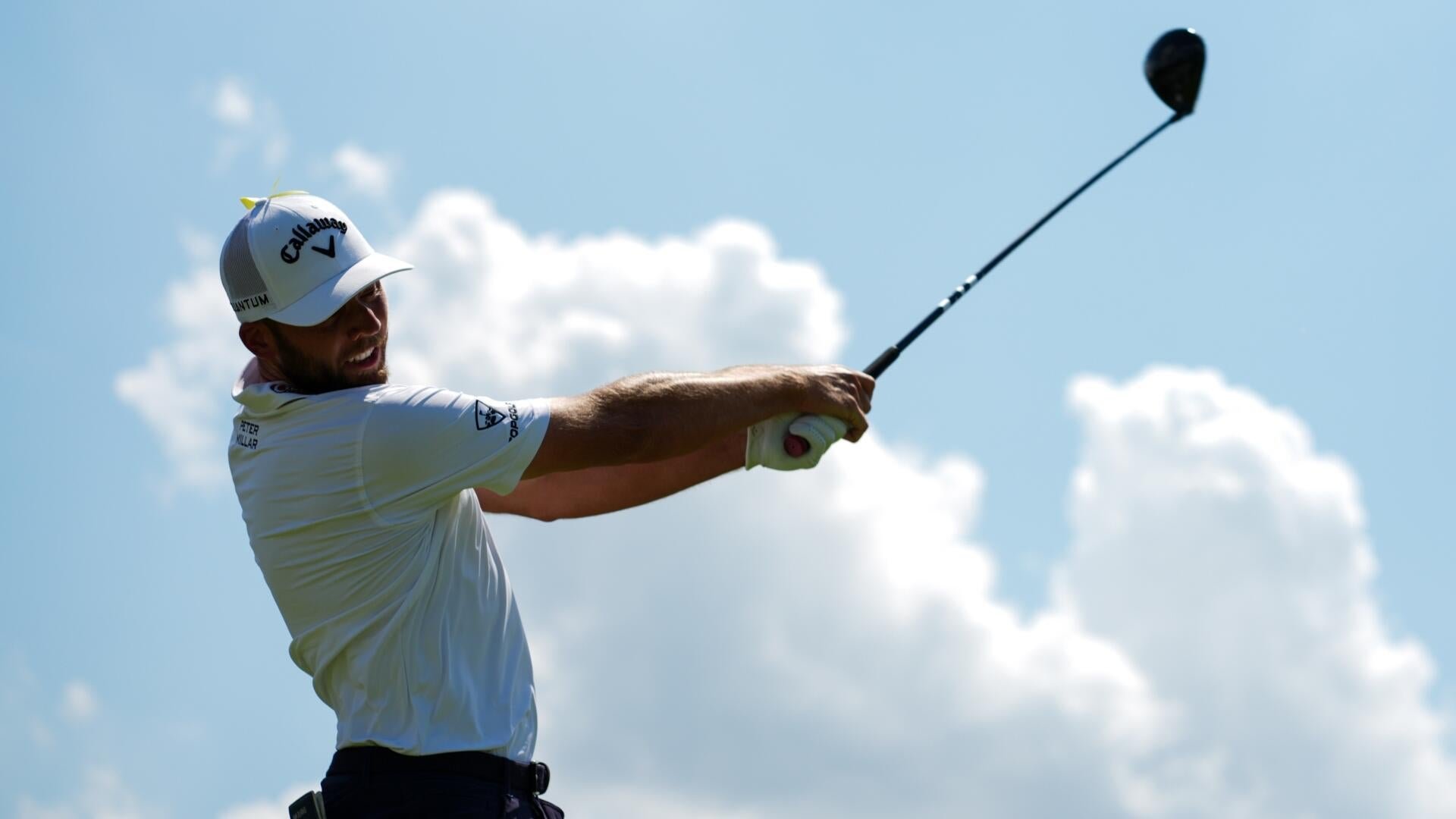 A golfer wearing a white Callaway cap and shirt completes a swing with a driver against a blue, cloudy sky.
