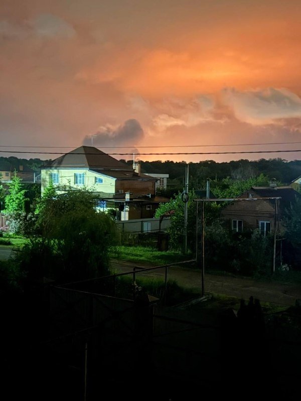 Houses, trees, and power lines are silhouetted beneath a dramatic orange and pink sunset sky with scattered clouds.