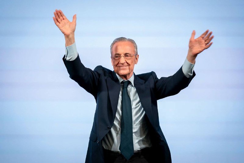 An older man wearing glasses, a dark suit, light blue shirt, and dark tie raises both arms with open palms against a light blue background.