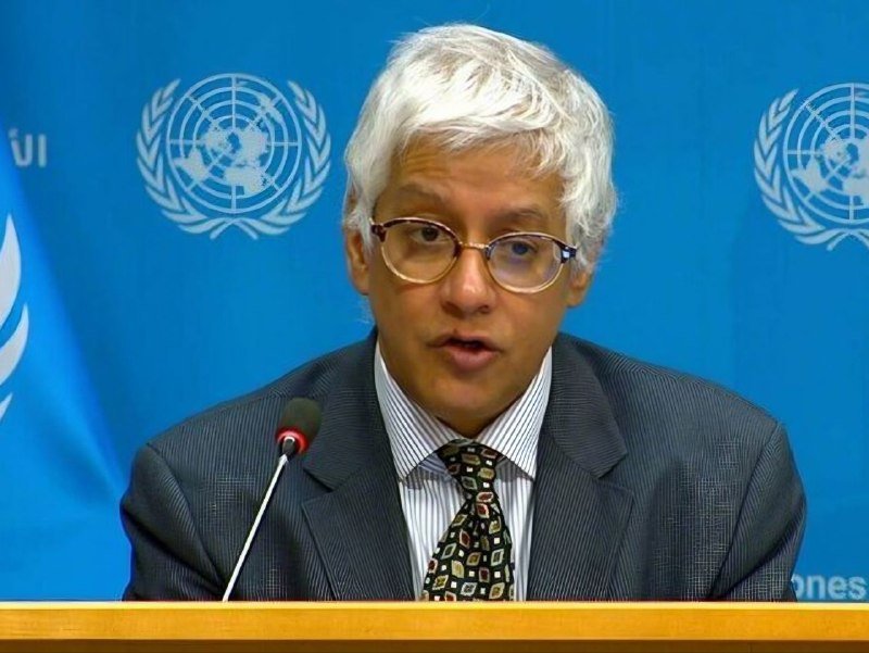 A white-haired man in glasses and a dark suit speaks at a podium microphone in front of a blue backdrop displaying the United Nations emblem.